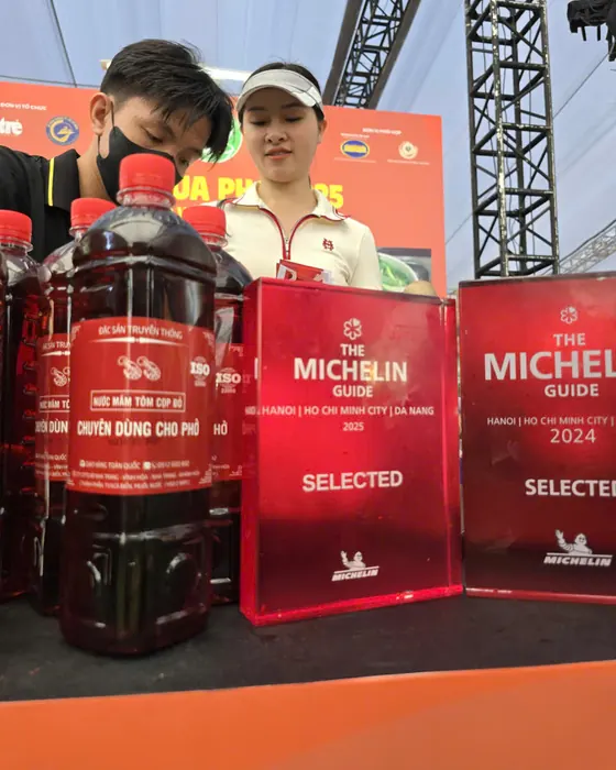 Hàng chai Tôm Cọp Đỏ đặt cạnh hai biển Michelin Guide Selected 2024 và 2025 của một gian quán phở